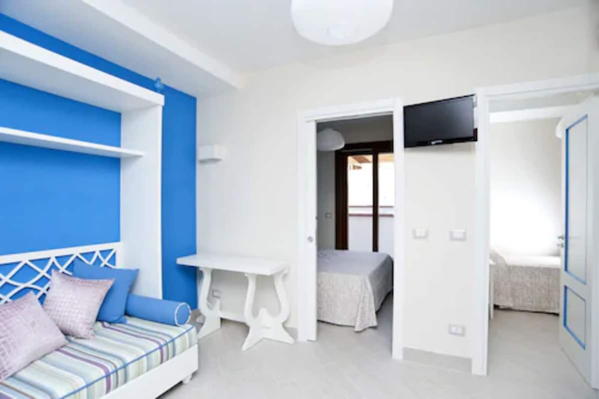 Ein modernes Schlafzimmer mit blauer Akzentwand, einem weißen Bett, einem Nachttisch und einem an der Wand montierten Fernseher.
