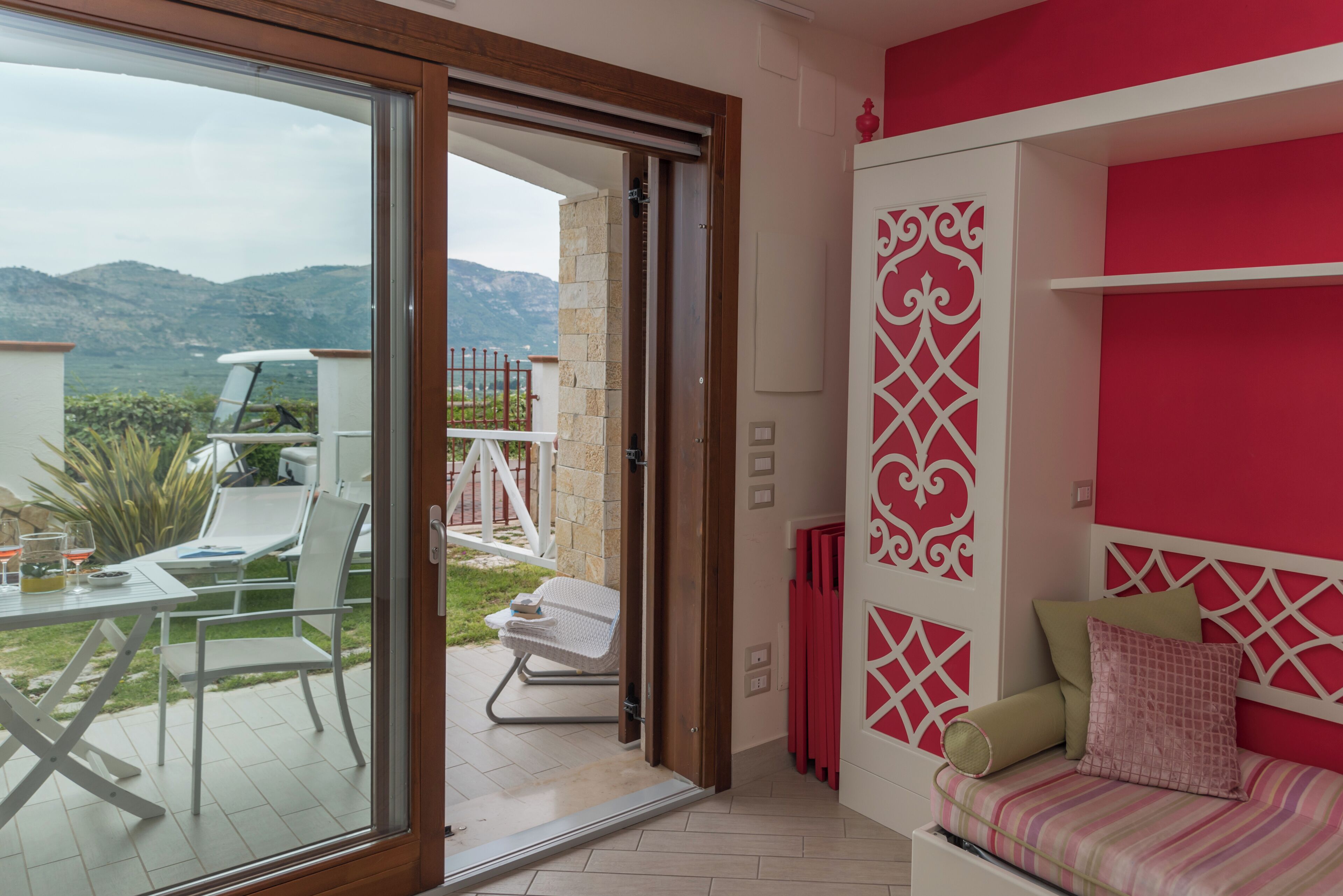 A room with a glass door leading to an outdoor area with a table and chairs, a bed with a red and white decorative headboard, and a view of the mountains.