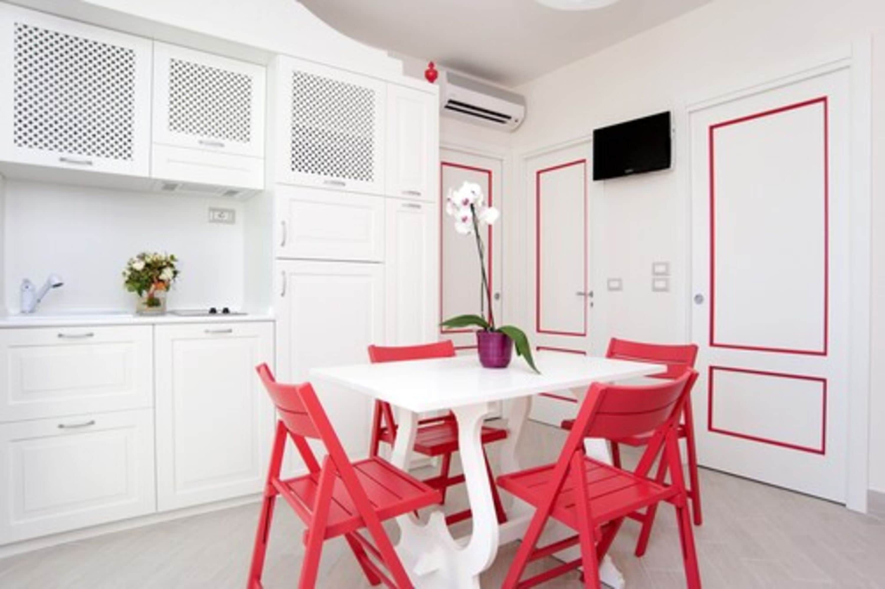 Una cocina moderna con armarios blancos, una mesa de comedor blanca con sillas rojas y una planta en maceta sobre la mesa.