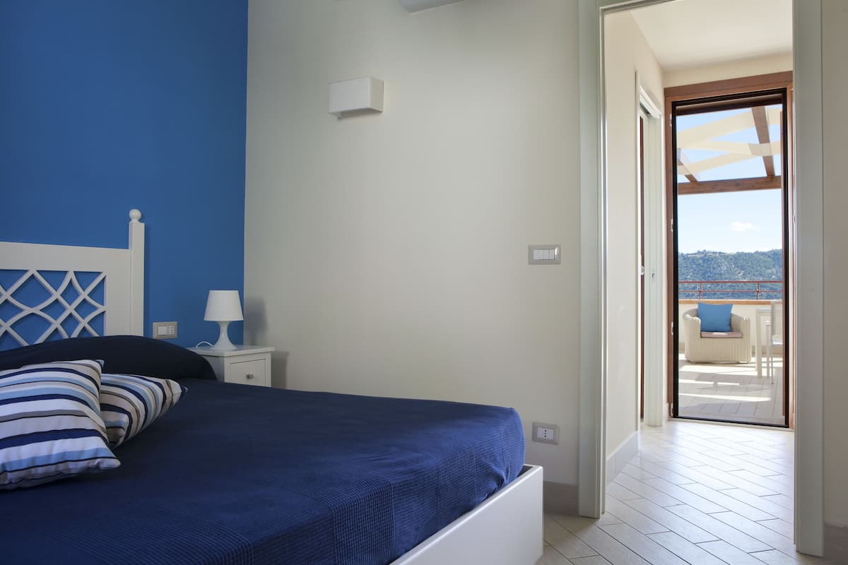 Ein Schlafzimmer mit Bett, Nachttisch, Lampe und Blick auf einen Balkon.