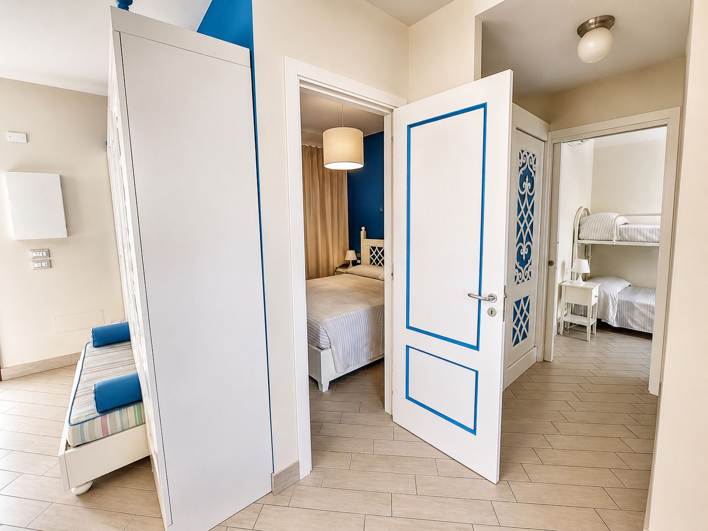 A hotel room with a bed, bedside table, lamp, and a view of another room with a bunk bed.