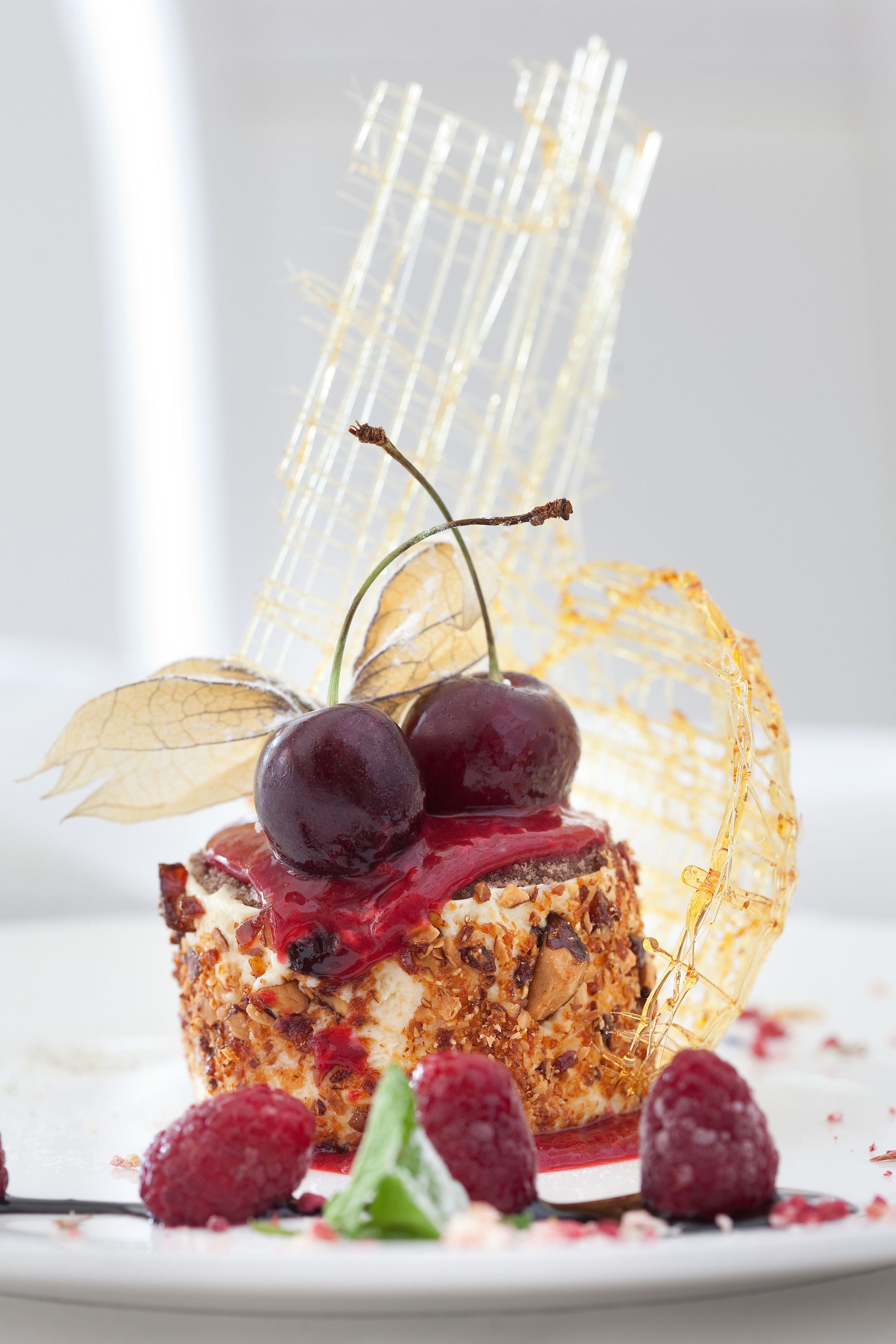 Een dessert met kersen, frambozen en een decoratief glaselement op een wit bord.