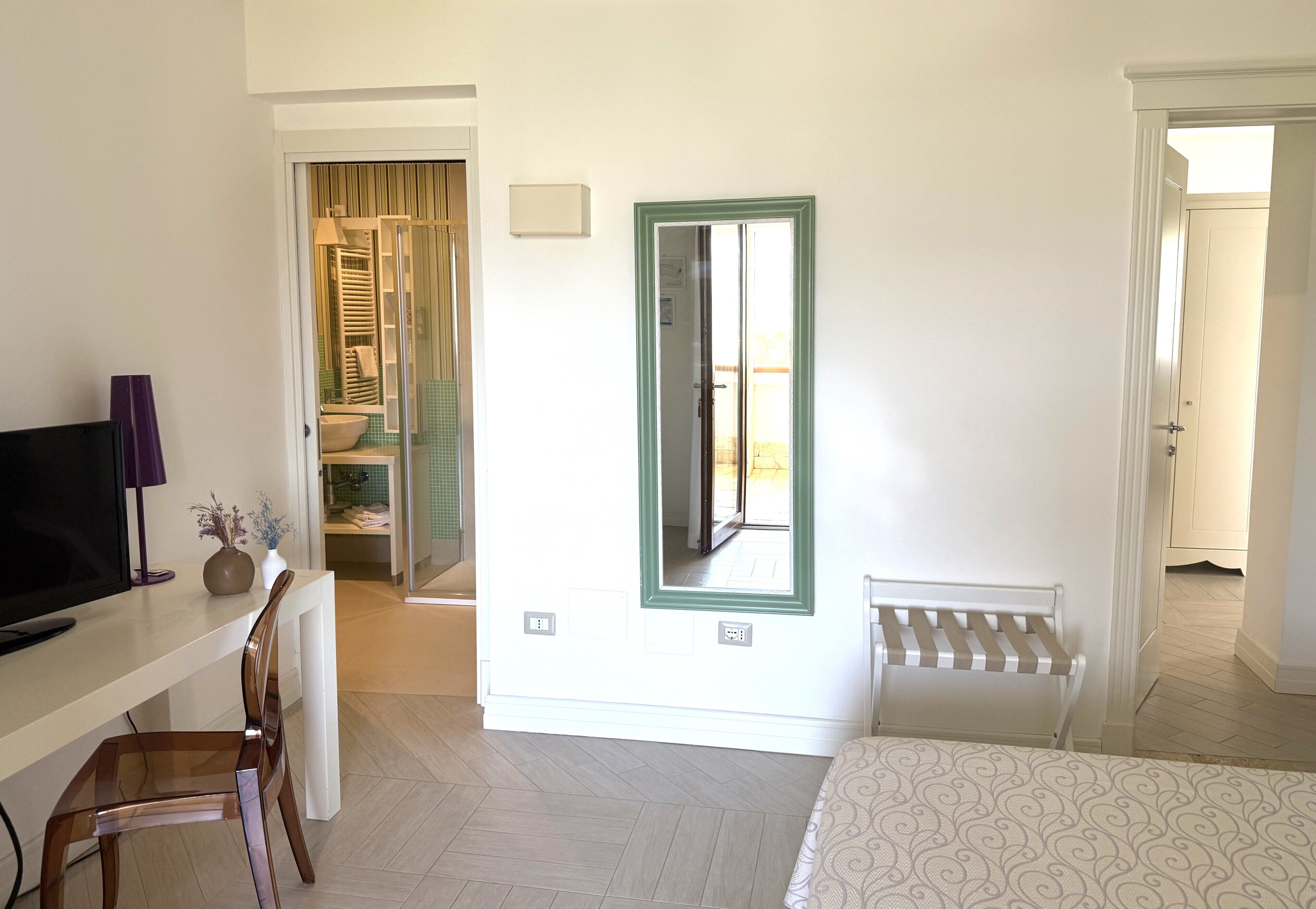 A hotel room with a bed, a desk with a TV, a chair, a radiator, and a window with a green frame.