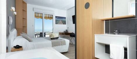 A modern hotel room with a large bed, a separate desk area, a bathroom with a large mirror, and a view of the sea.