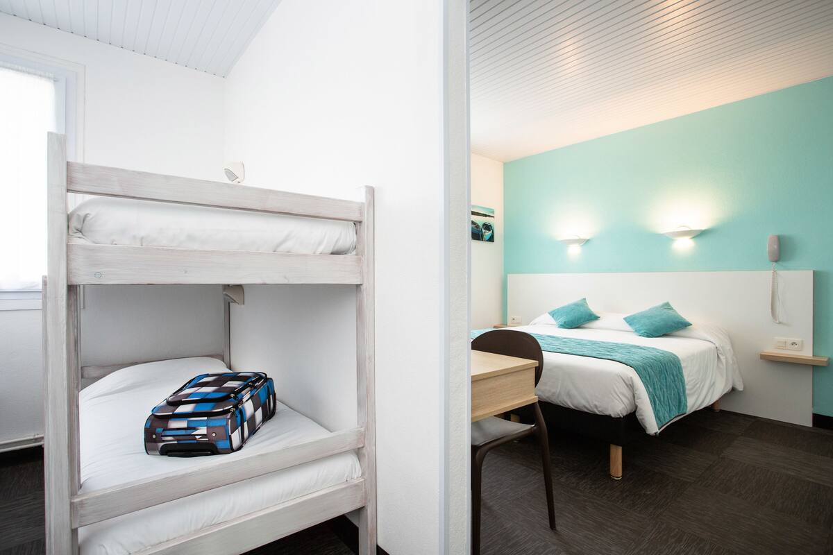 Un lit superposé avec une valise posée sur le lit du bas et un lit simple avec un bureau et une chaise dans la pièce adjacente.