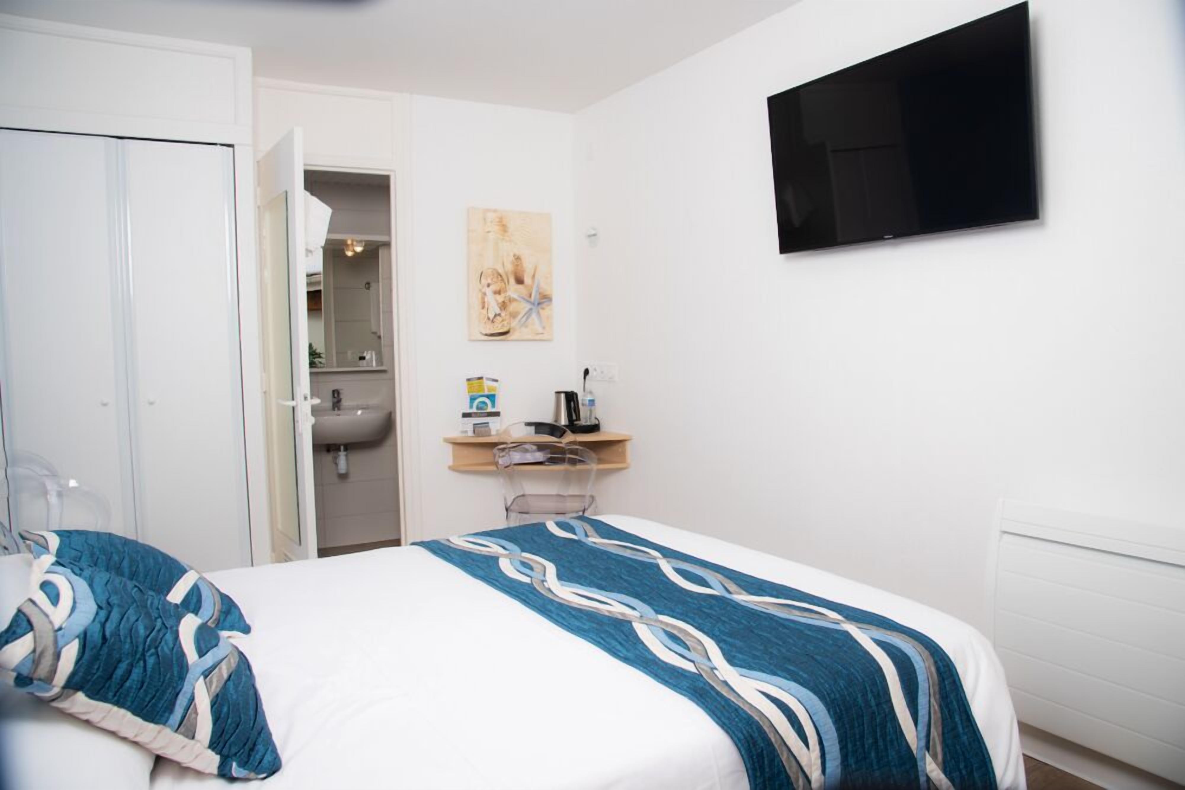 A hotel room with a bed, a bedside table with a lamp, a TV mounted on the wall, and a bathroom visible through an open door.