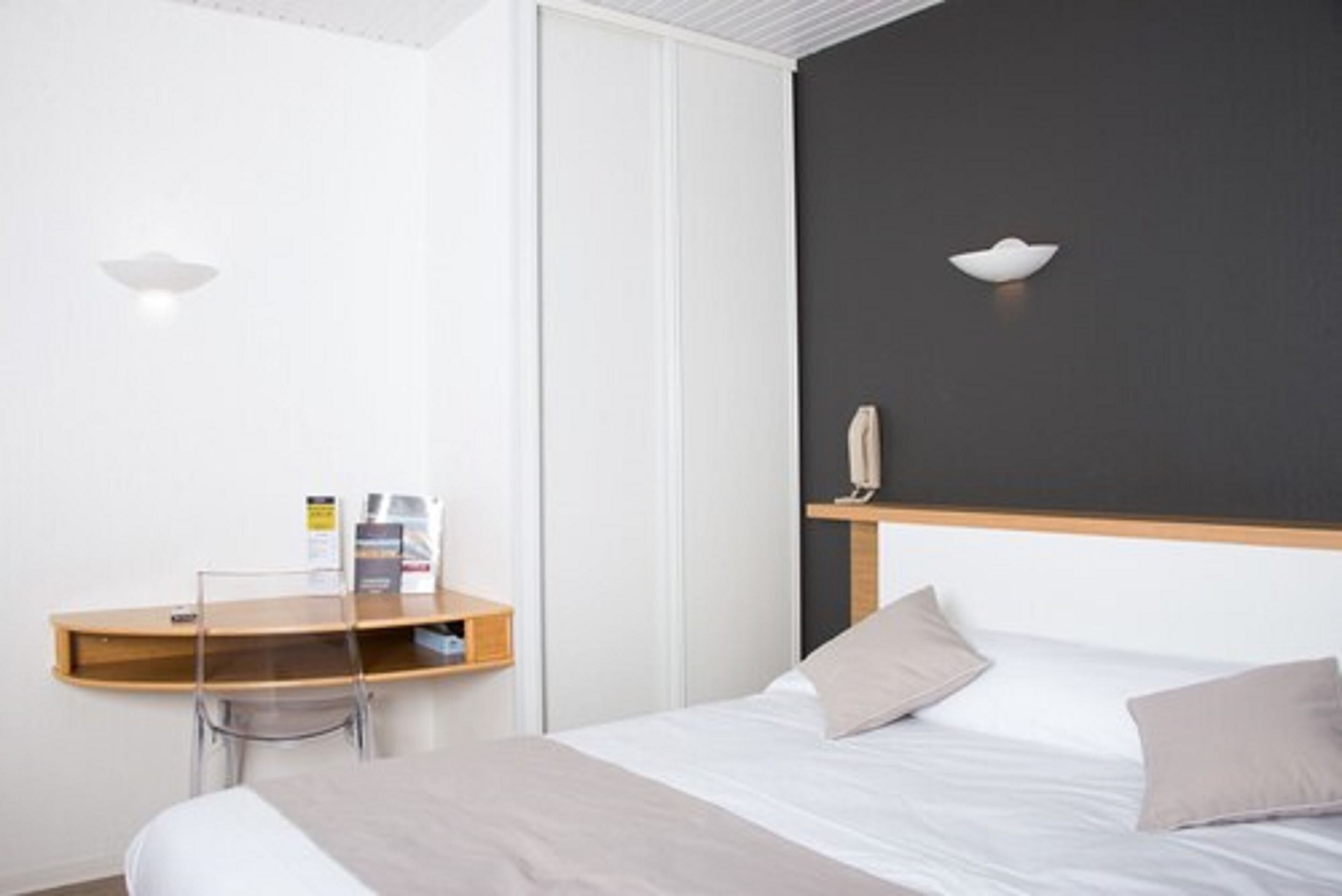 A hotel room with a bed, a desk with a chair, a white wardrobe, and a dark accent wall.
