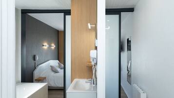 Habitación de hotel moderna con cama, mesita de noche y baño con lavamanos y espejo.