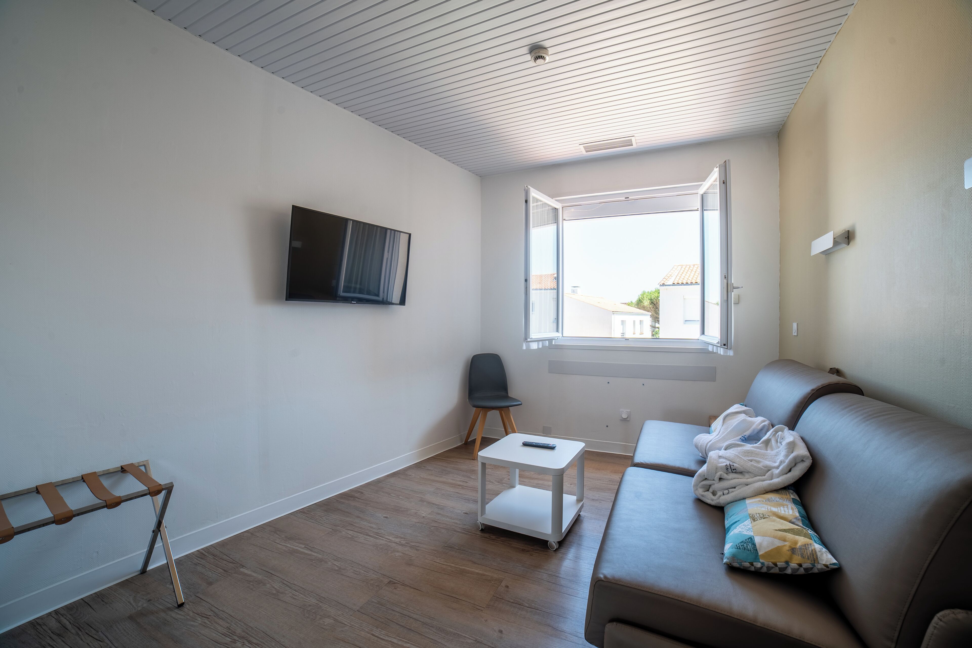 Un salon moderne avec un canapé en cuir marron, une petite table blanche et une chaise noire. Une fenêtre laisse entrer la lumière naturelle, et un téléviseur à écran plat est fixé au mur.