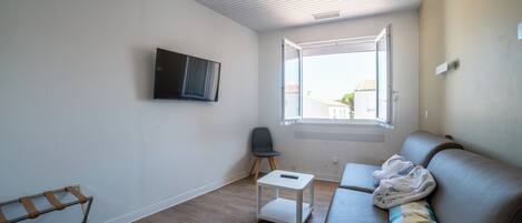 Un salon moderne avec un canapé en cuir marron, une petite table blanche et une chaise noire. Une fenêtre laisse entrer la lumière naturelle, et un téléviseur à écran plat est fixé au mur.