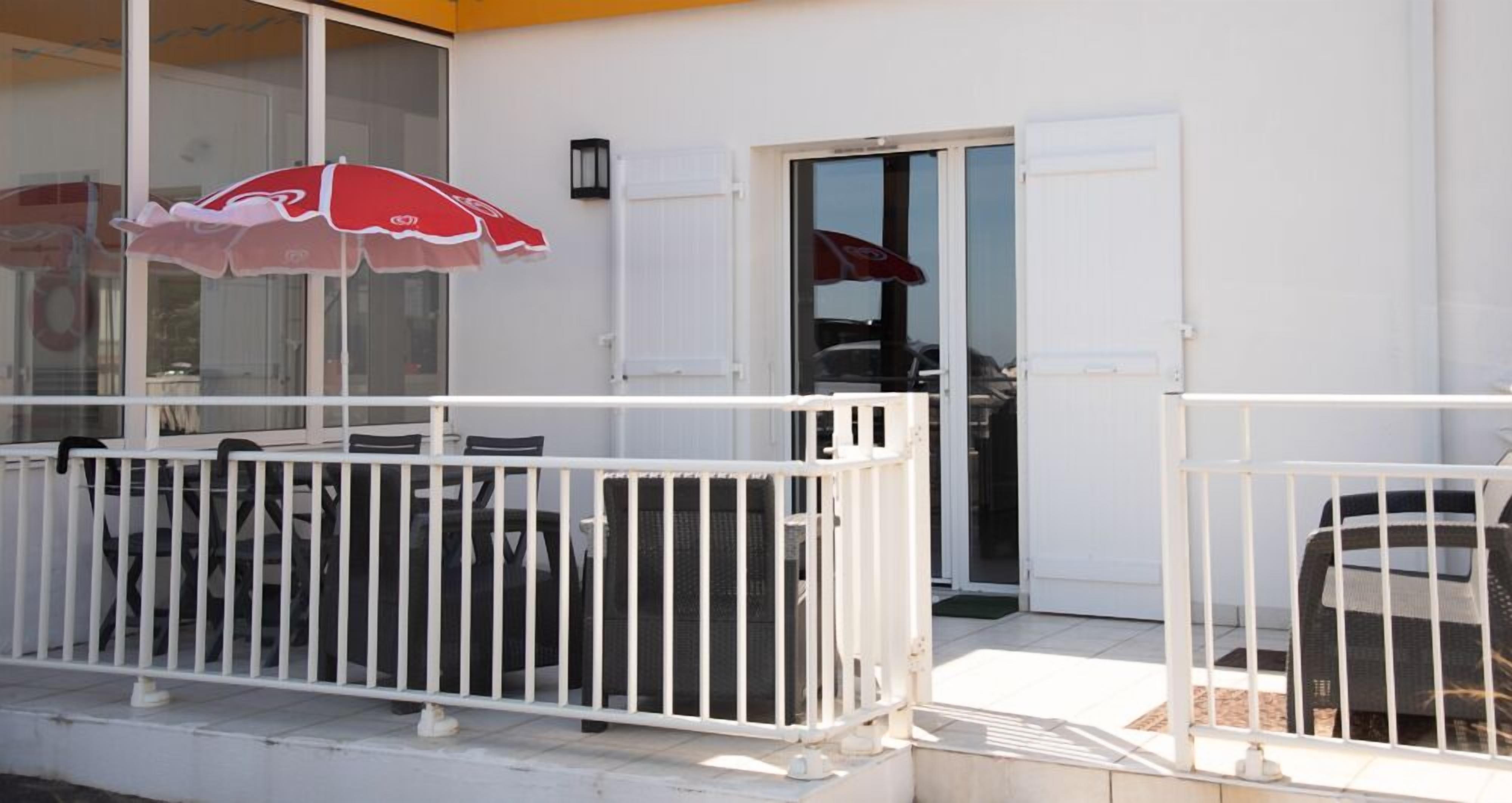 Un balcone bianco con ringhiera bianca, un ombrellone rosso e una porta che dà su un ambiente interno.