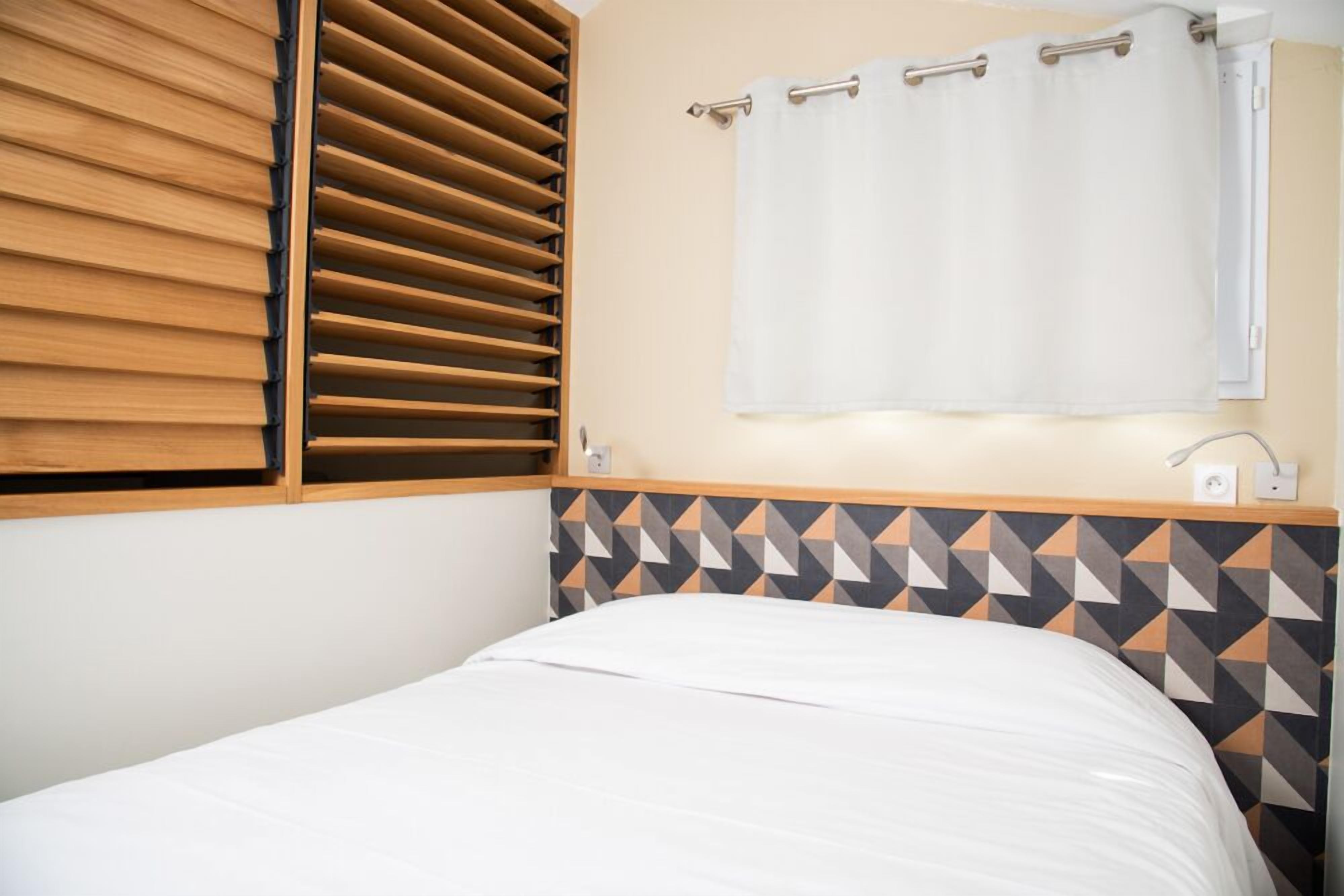 Un dormitorio con cama, persianas de madera, pared con diseño geométrico, cortina blanca e interruptor de luz en la pared.