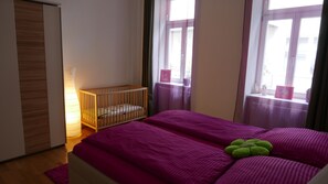 A room with a crib, a bed with a purple bedspread, and a window with curtains.