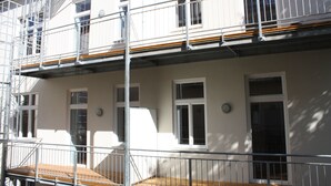 Un immeuble à plusieurs étages, aux murs extérieurs blancs, doté de nombreuses fenêtres et d’un balcon avec une rambarde en métal.
