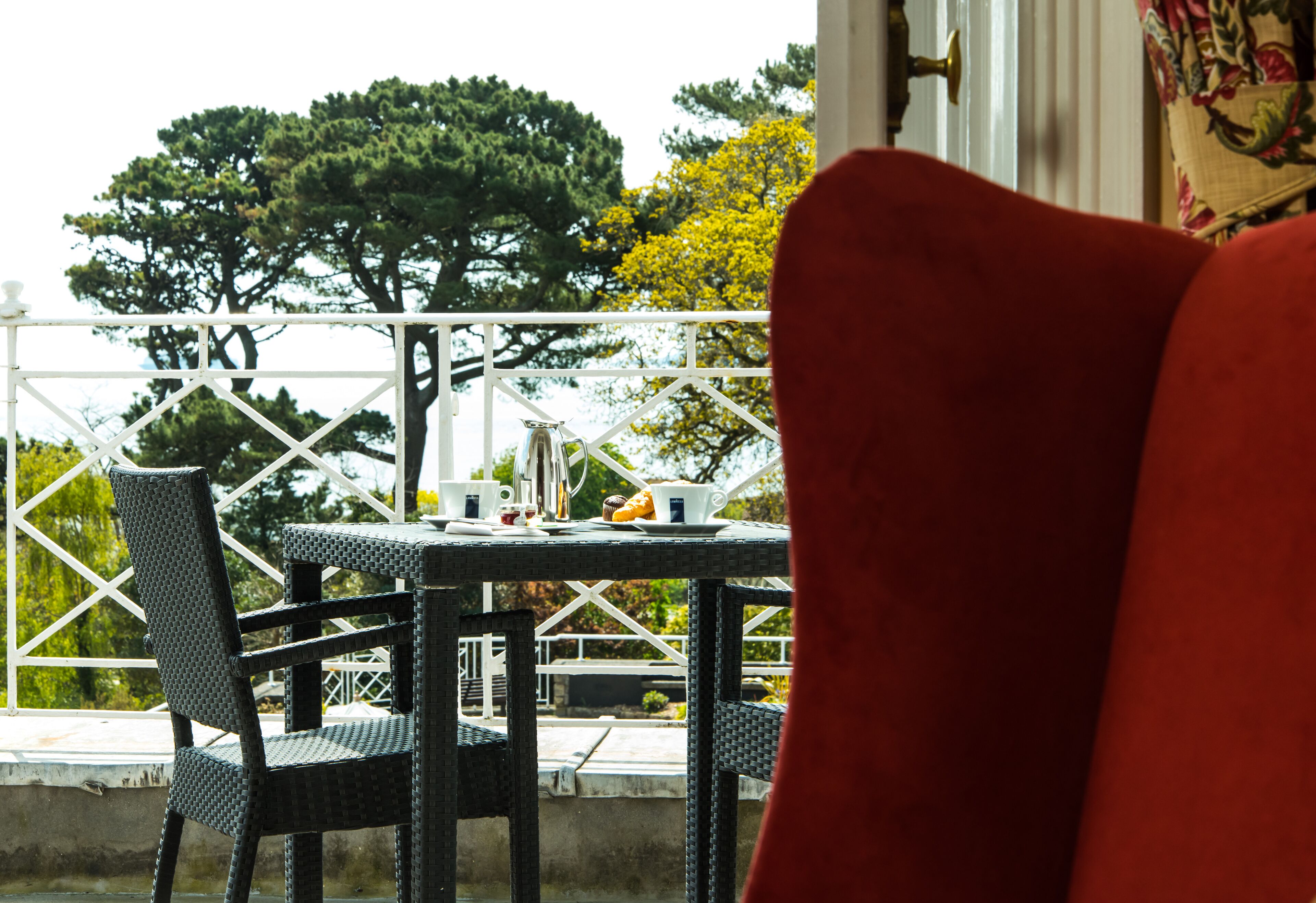 Een balkon met rieten stoelen en een tafel voor twee, met uitzicht op een tuin met bomen en een witte hekken.