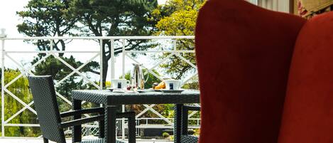 A balcony with wicker chairs and a table set for two, overlooking a garden with trees and a white fence.