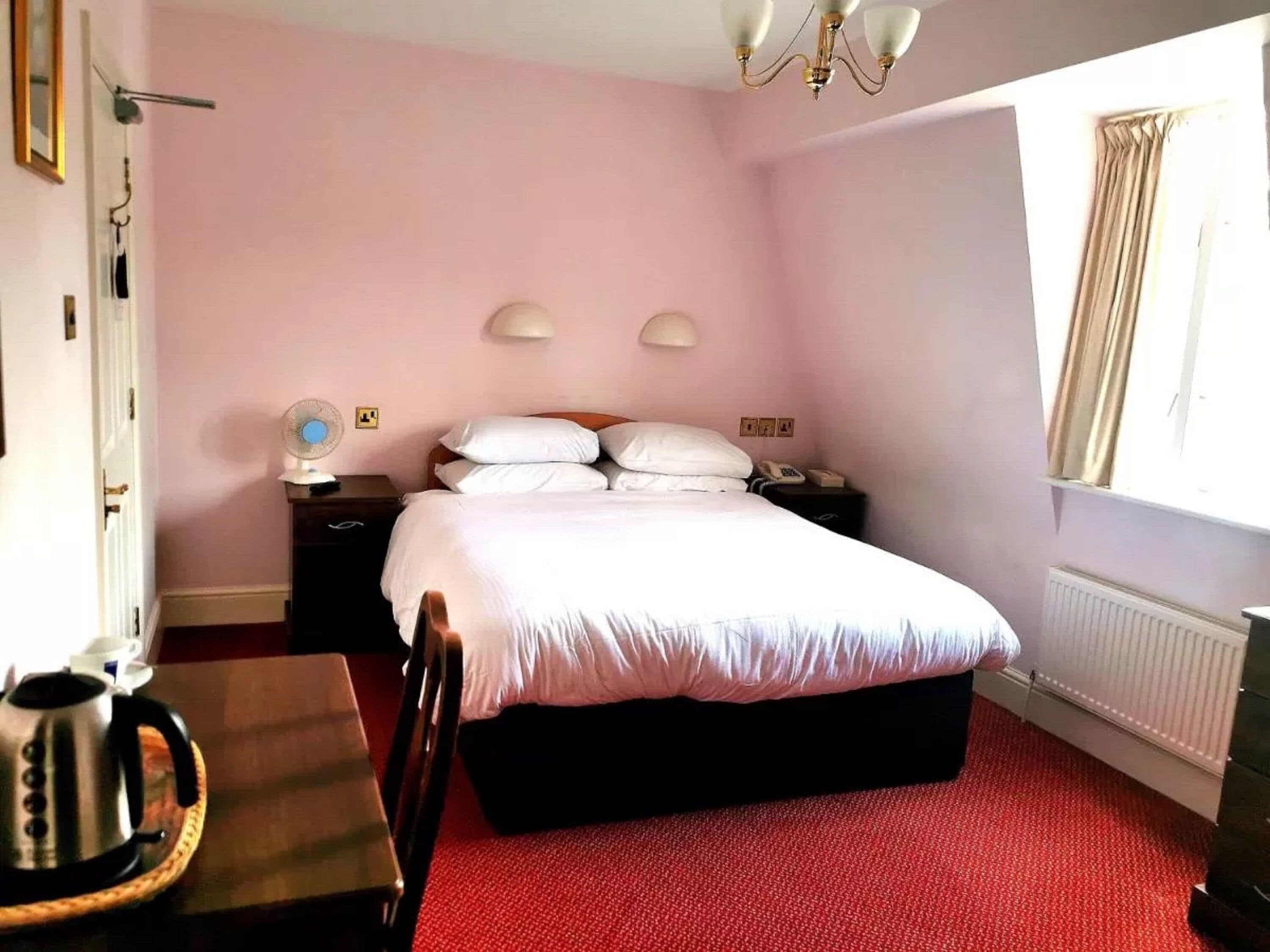 Habitación de hotel con cama, mesitas de noche, escritorio con hervidor, silla, radiador y ventana con cortinas.