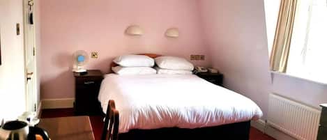 Habitación de hotel con cama, mesitas de noche, escritorio con hervidor, silla, radiador y ventana con cortinas.
