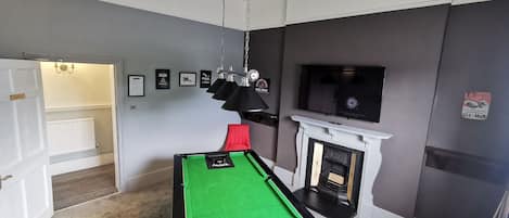 Une table de billard dans une pièce avec une cheminée, un téléviseur fixé au mur et des tableaux encadrés accrochés au mur.