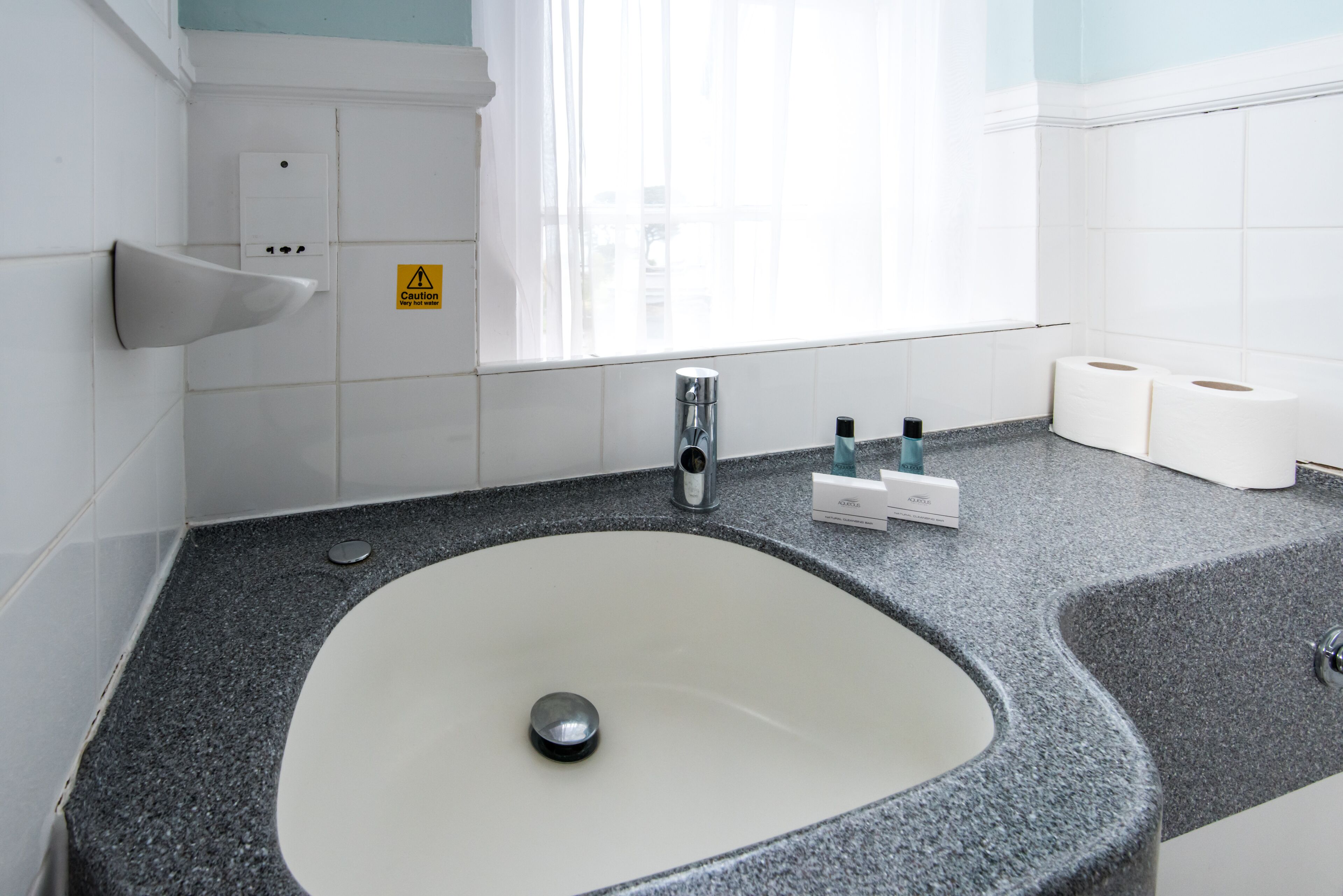 A bathroom with a granite countertop, a white sink, a faucet, a caution sign, and toilet paper.