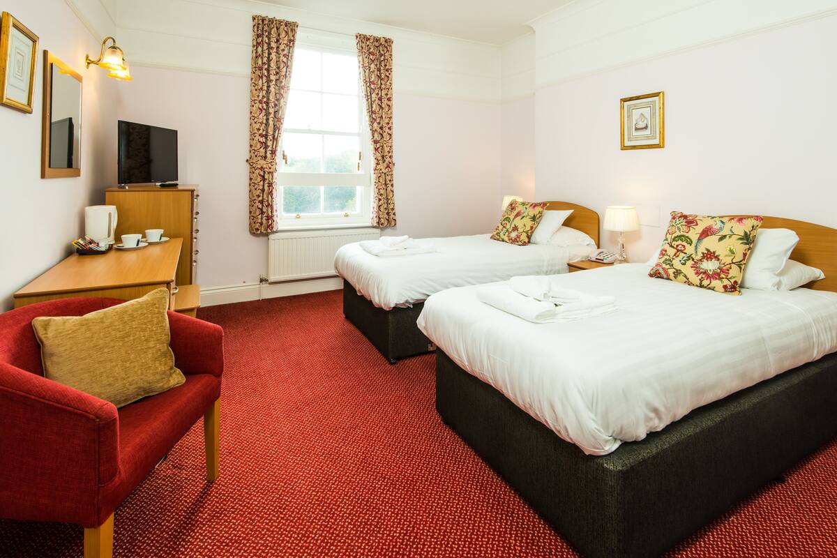 A hotel room with two beds, a desk with a TV, a red armchair, and a window with curtains.