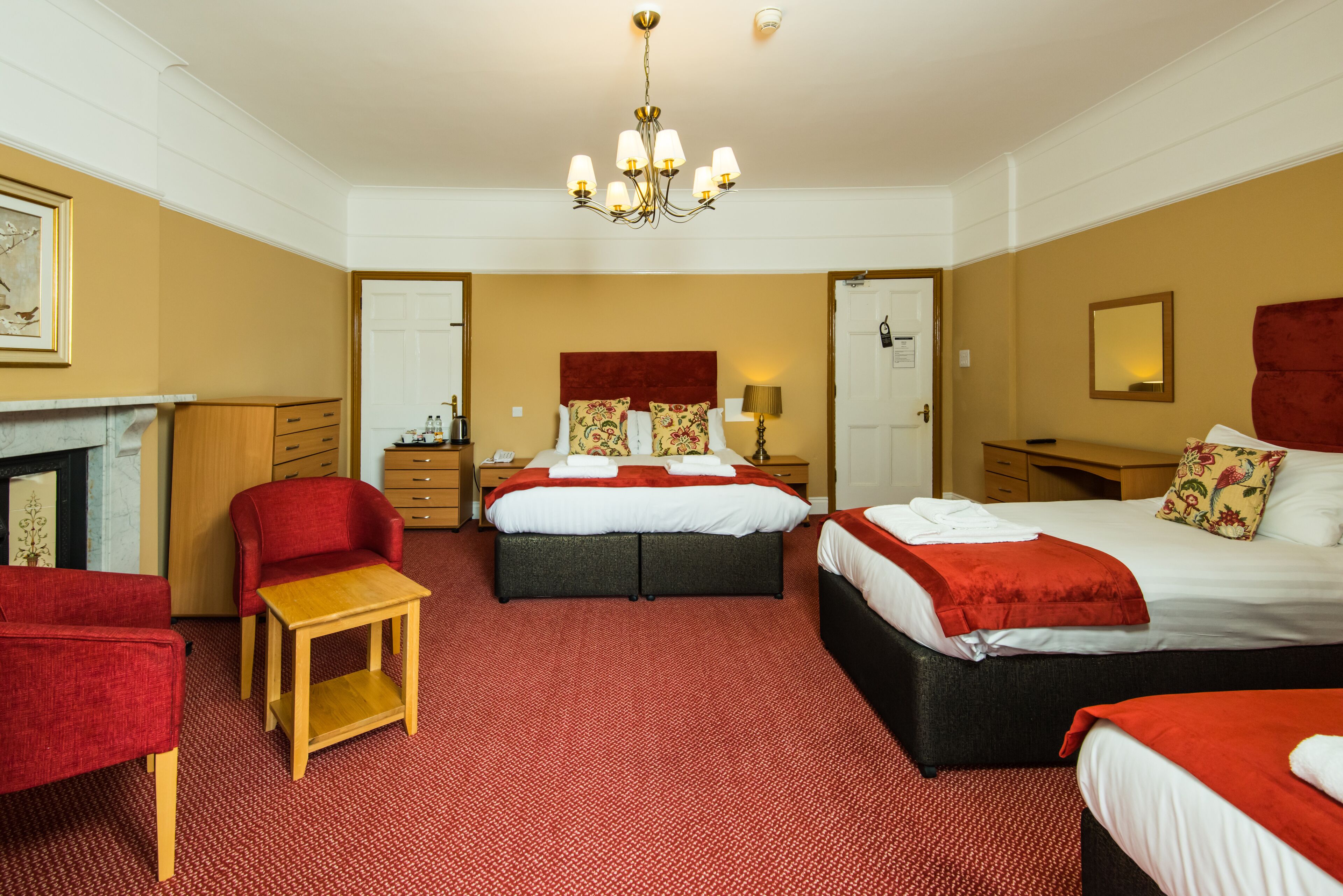 Habitación de hotel con dos camas, alfombra roja, mesita de madera, sillón rojo, candelabro y chimenea.