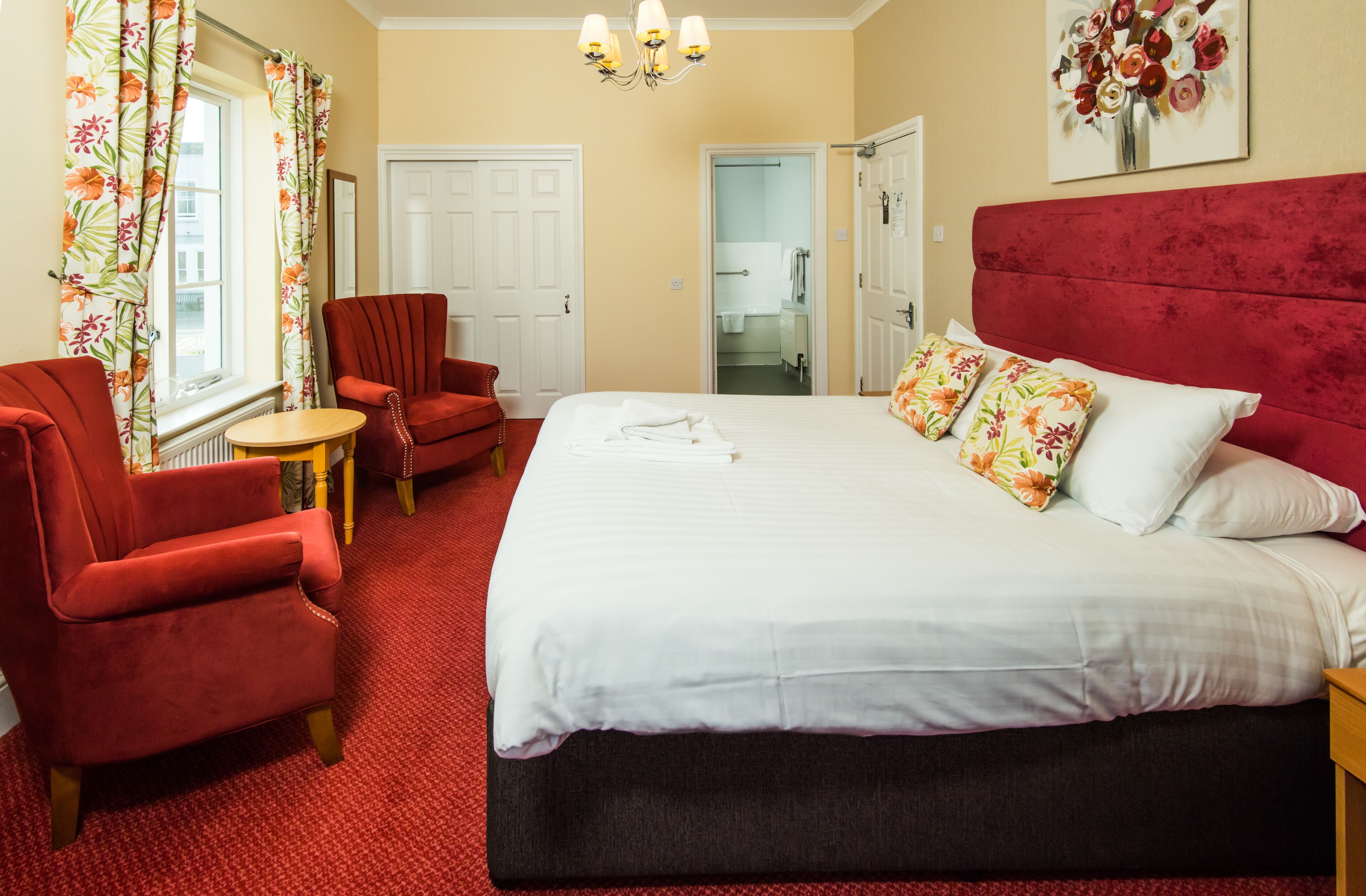 A hotel room with a large bed, red armchairs, a small round table, and a red headboard.