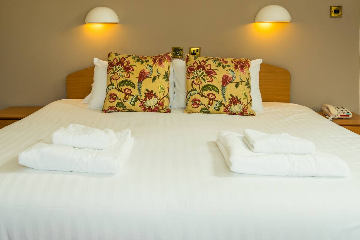 A neatly made double bed with floral pillows, two bedside lamps, a phone, and folded towels on a clean, white bedspread.