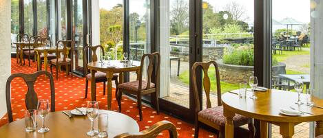 A restaurant with round tables and chairs, large windows, and a red carpet.