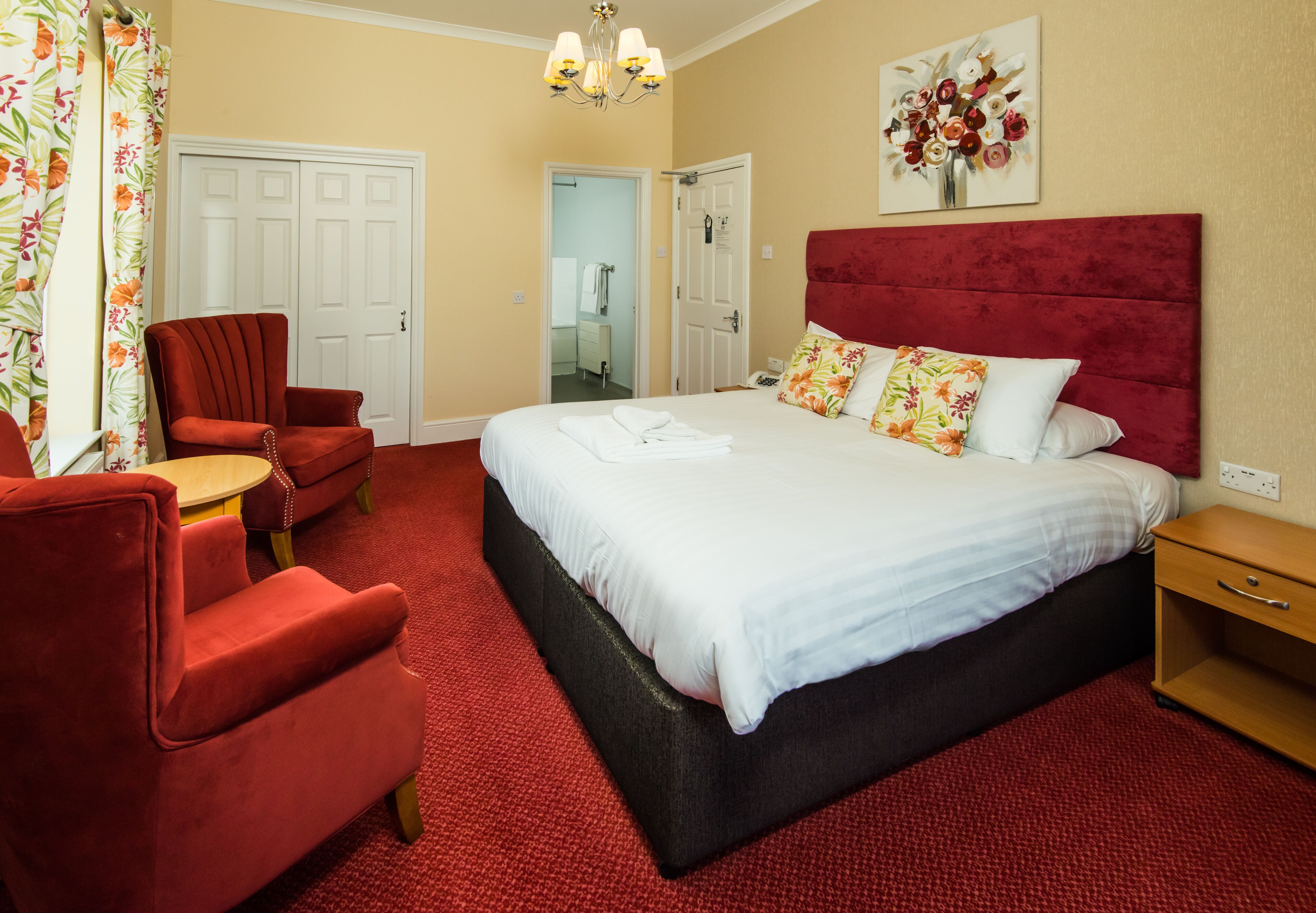 Una habitación de hotel con una cama grande, un sillón rojo, una mesita de noche de madera y un cuadro floral en la pared.