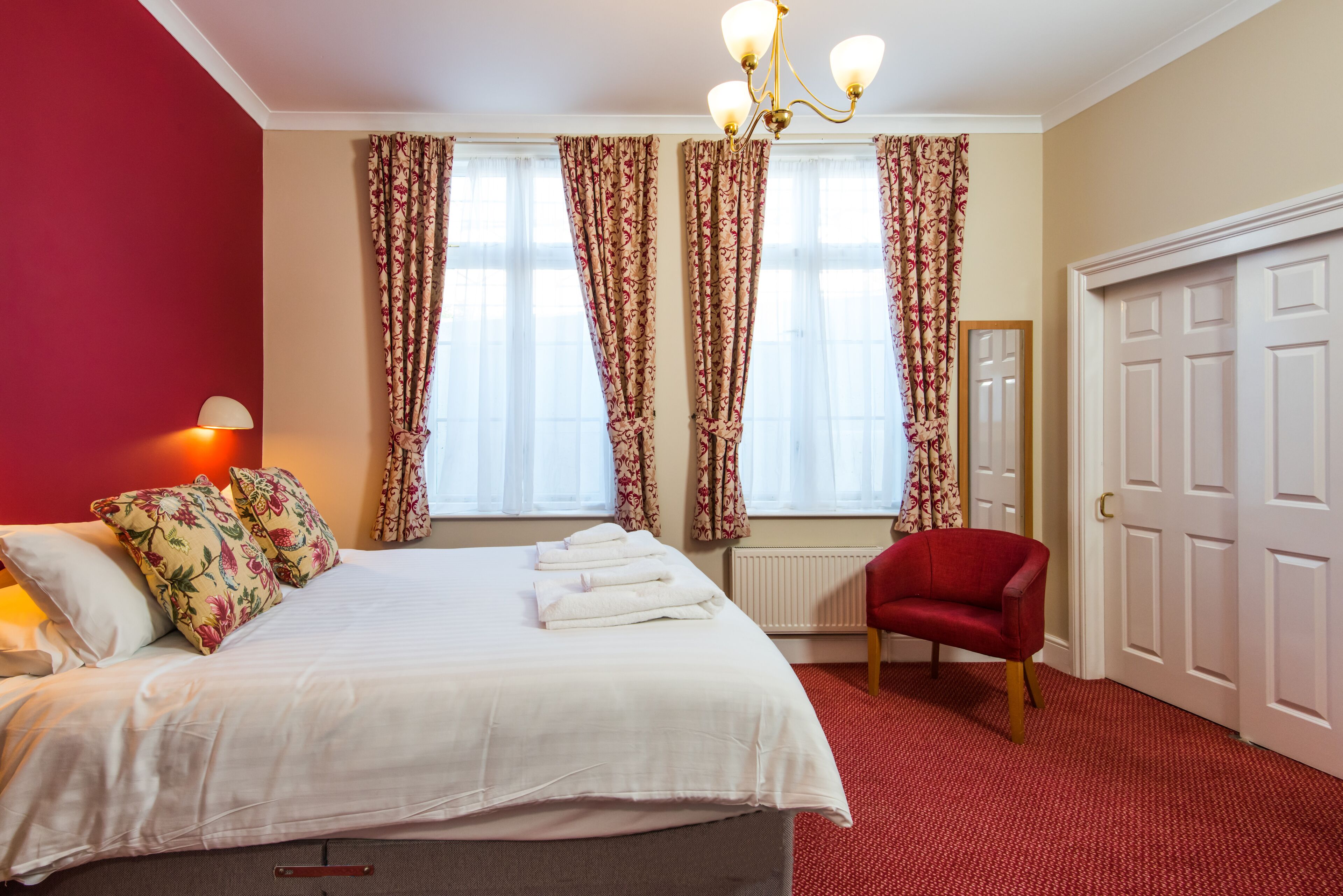 A hotel room with a bed, a red armchair, a standing lamp, and a large window with curtains.