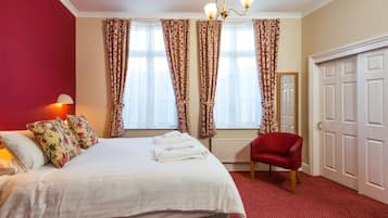 A hotel room with a bed, a red armchair, a standing lamp, and a large window with curtains.