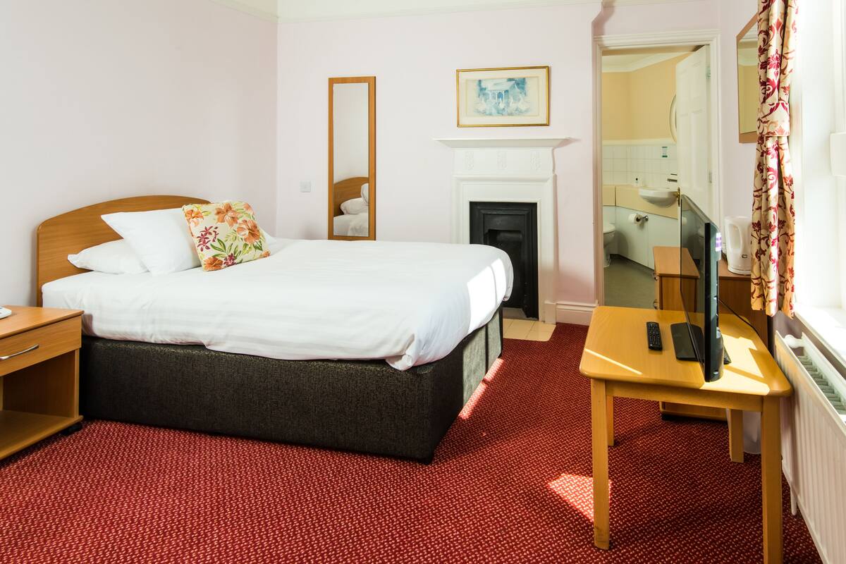 A hotel room with a bed, a bedside table with a TV, a fireplace, and a bathroom visible through an open door.
