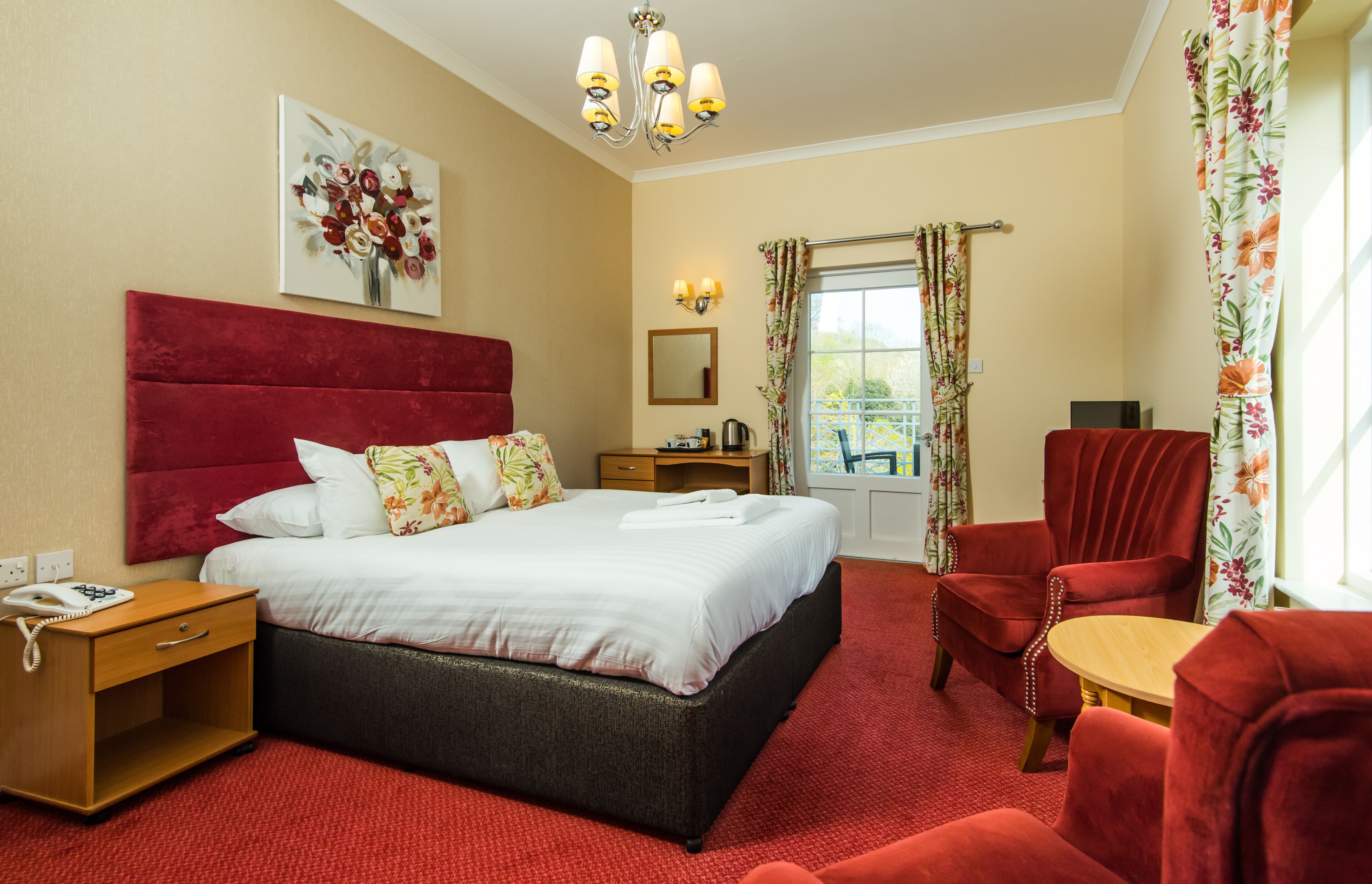 Ein Hotelzimmer mit einem großen Bett, einem rot gepolsterten Sessel, einem Holznachttisch, einem kleinen Tisch und einem Fenster mit Vorhängen.