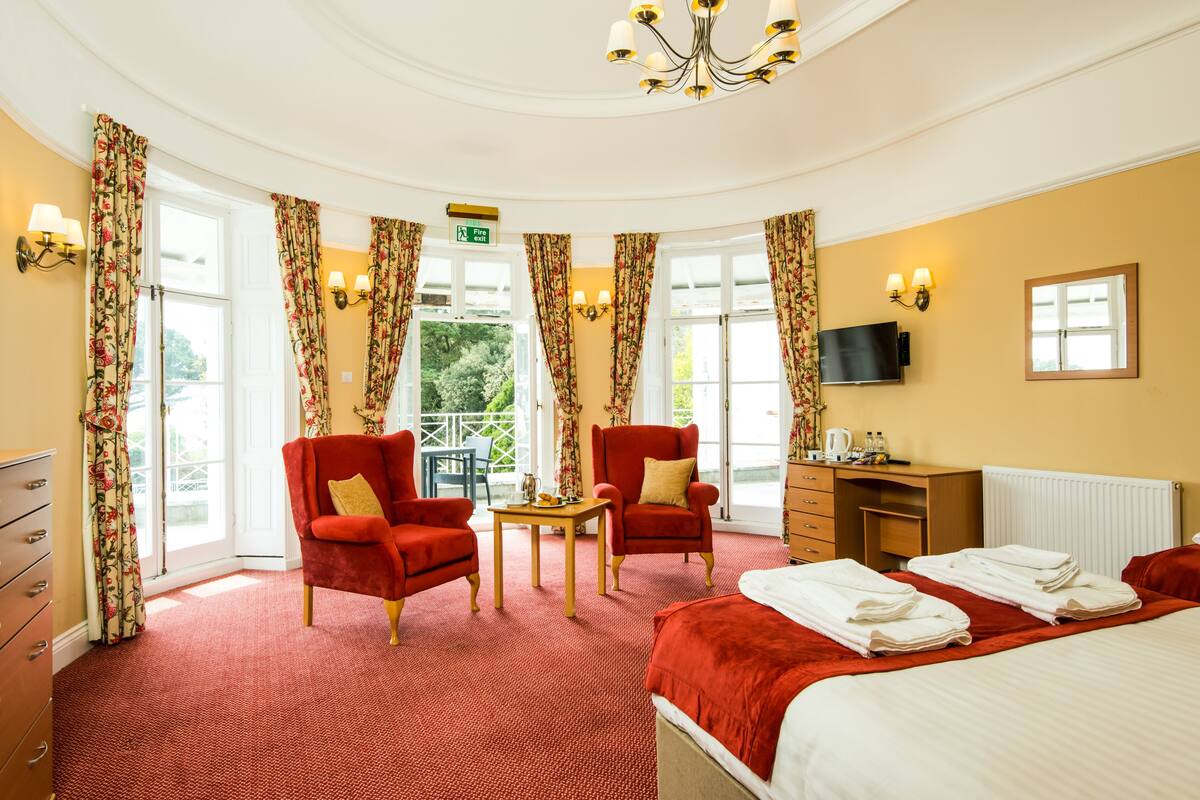 A hotel room with a red carpet, two red armchairs, a bed with white linens, a desk with a flat-screen TV, and a chandelier.