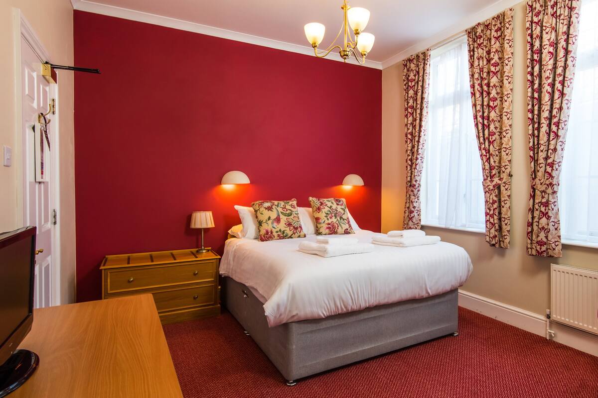 A hotel room with a red wall, a bed with white linens, two bedside lamps, a wooden dresser, a red armchair, and floral curtains.