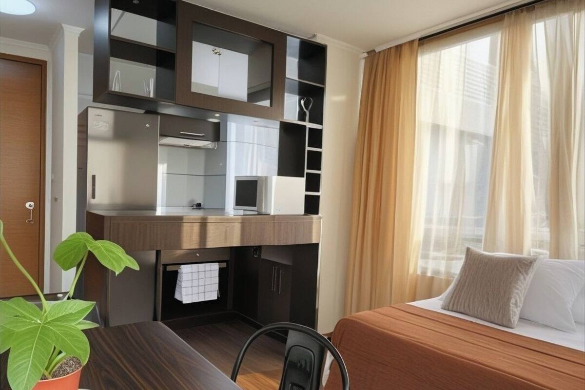 Ein kompaktes Hotelzimmer mit einem Holztisch, einem Bett mit beige farbenem Bettbezug, einem eingebauten Kleiderschrank mit Spiegeln und einer Topfpflanze.