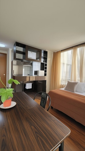 Une chambre d’hôtel compacte comprenant une table à manger en bois, un lit avec une couvre-lit beige, une armoire intégrée avec des miroirs et une plante en pot.