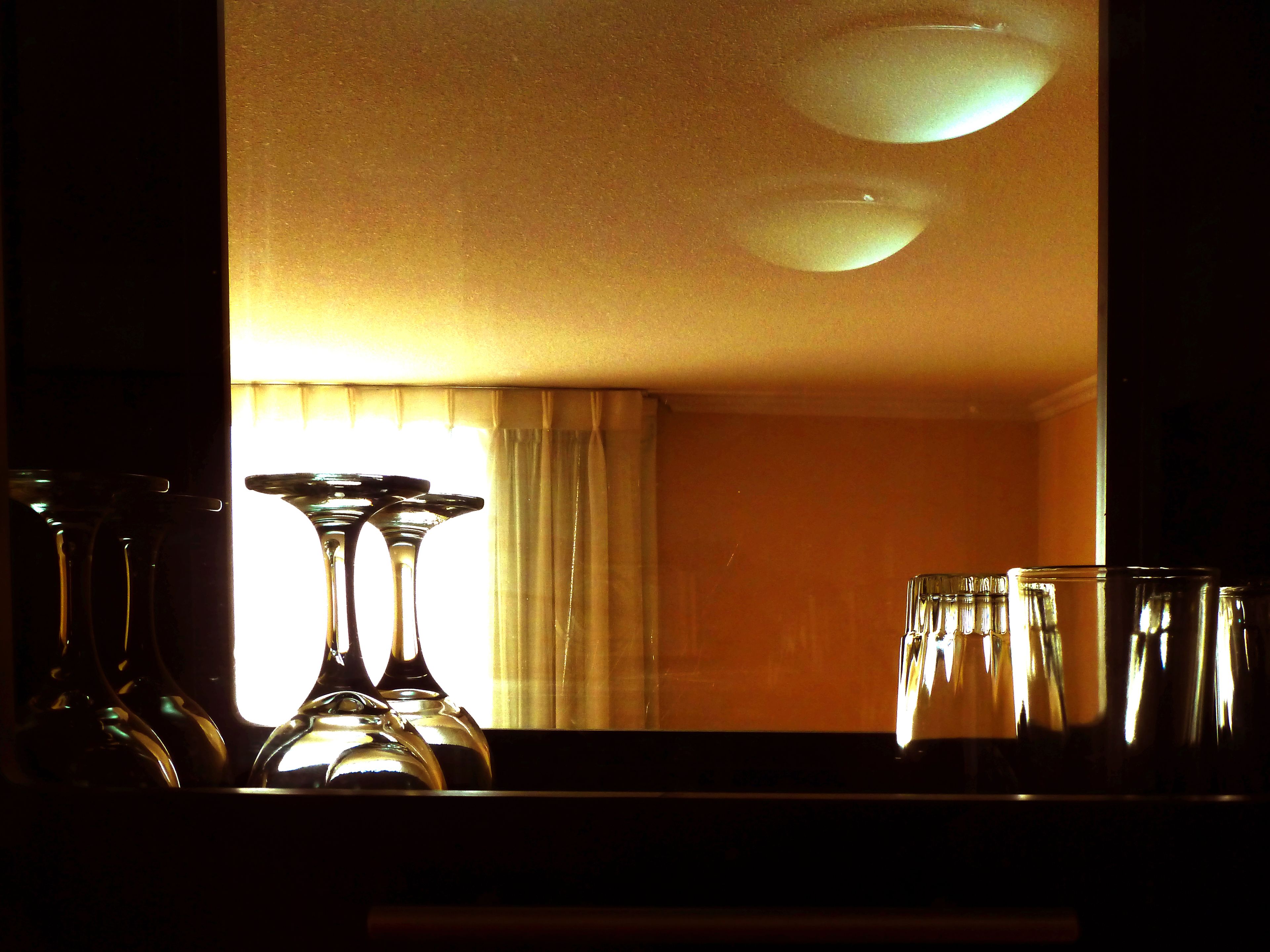 Ett rum med en stor spegel som reflekterar ett fönster med gardiner, en lampa och flera glas på ett bord.