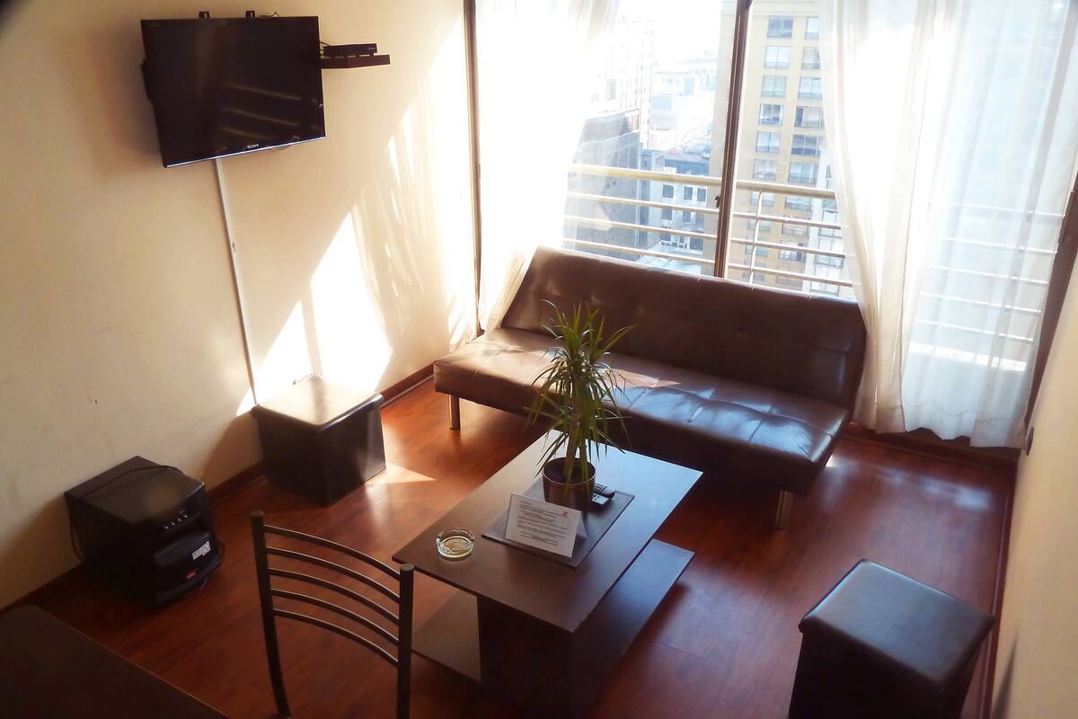 Ein Wohnzimmer mit einer Couch, einem Couchtisch mit einer Pflanze, einem an der Wand montierten Fernseher und einem Fenster mit Vorhängen.