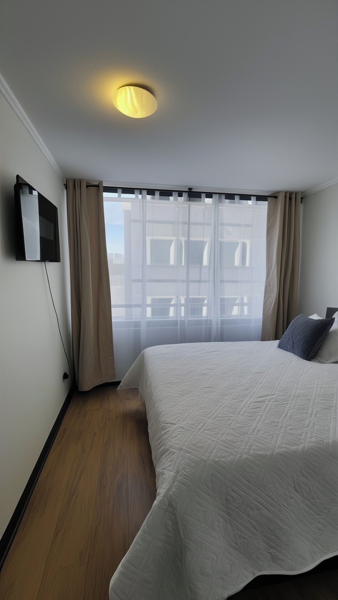 A hotel room with a bed, a TV mounted on the wall, a window with curtains, and a ceiling light.