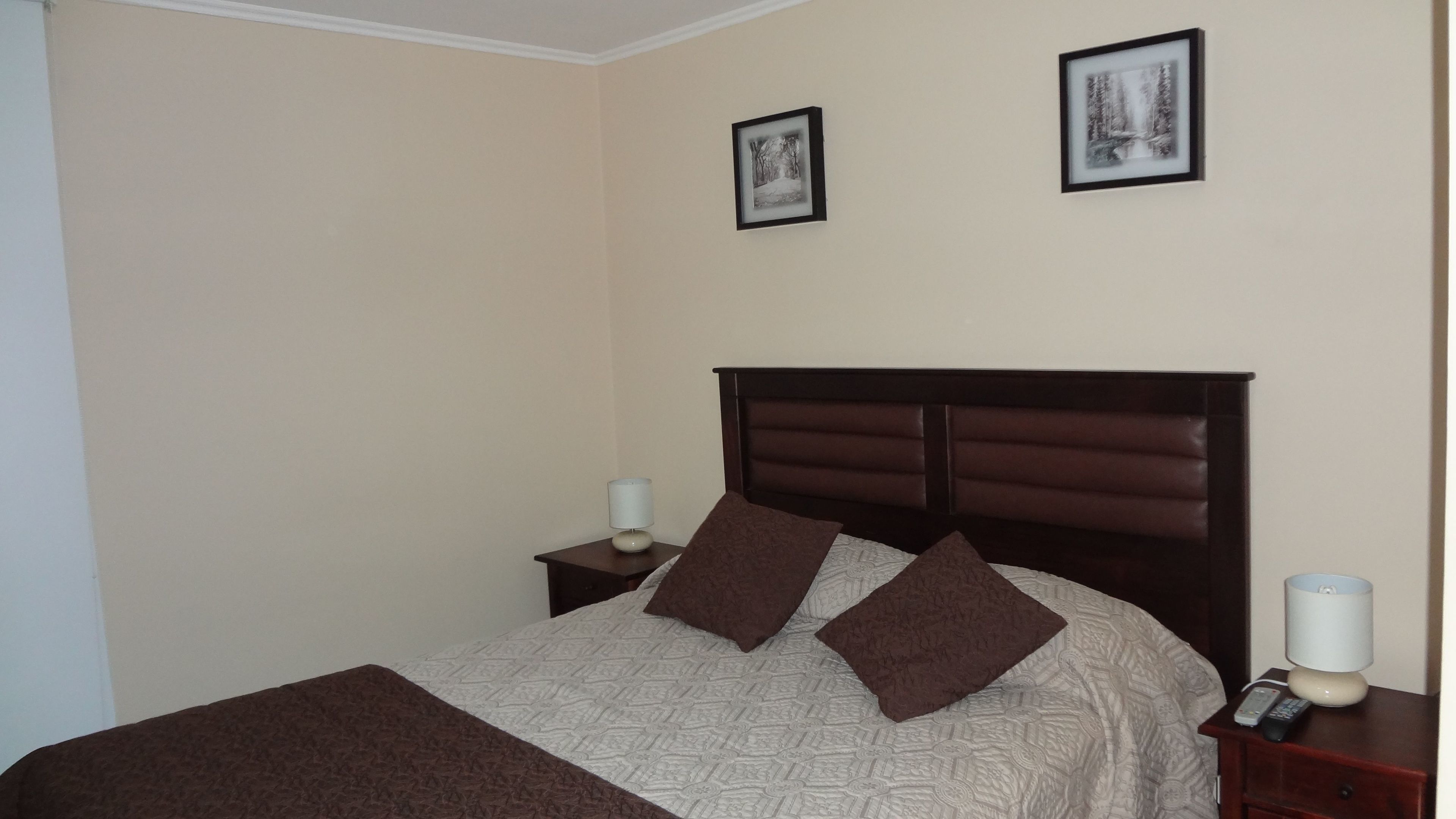 Una cama bien hecha con un edredón estampado y dos almohadas, una mesita de noche con una lámpara y un control remoto, y dos cuadros enmarcados en la pared.