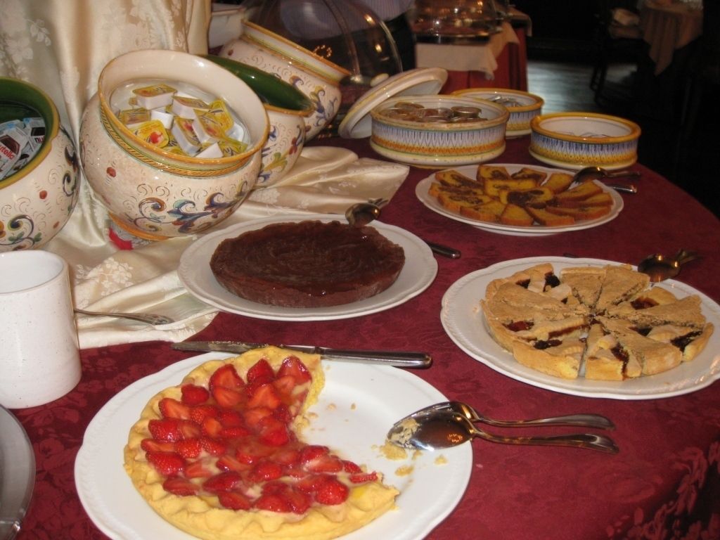 Un tavolo con diversi piatti, tra cui una crostata di fragole, una torta al cioccolato e una torta di frutta. Si vedono anche ciotole decorative e una tazza bianca.