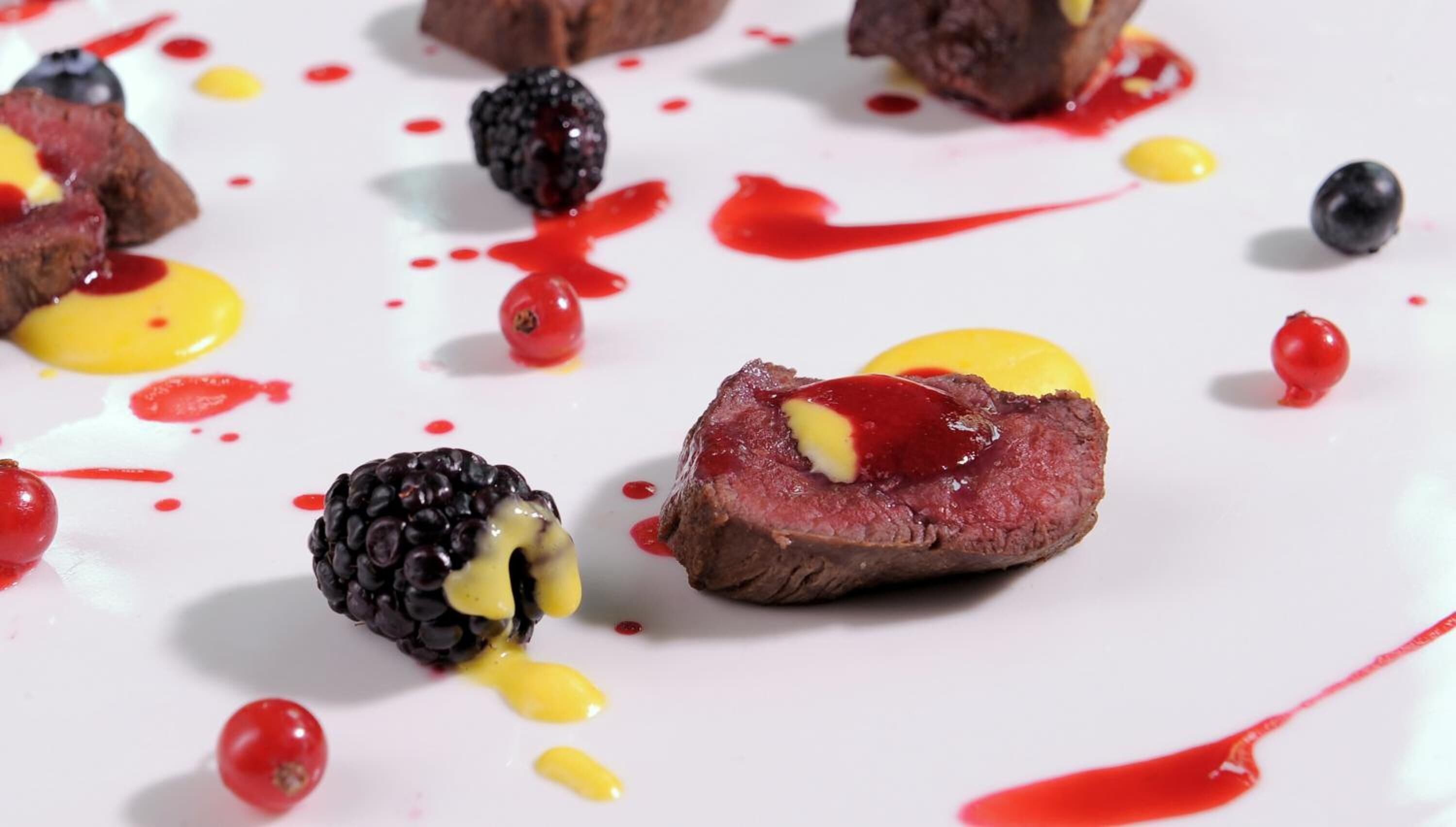 Een dessertborden met vleesschijf, gegarneerd met bramen en gele saus, omringd door bessen en rode saus.