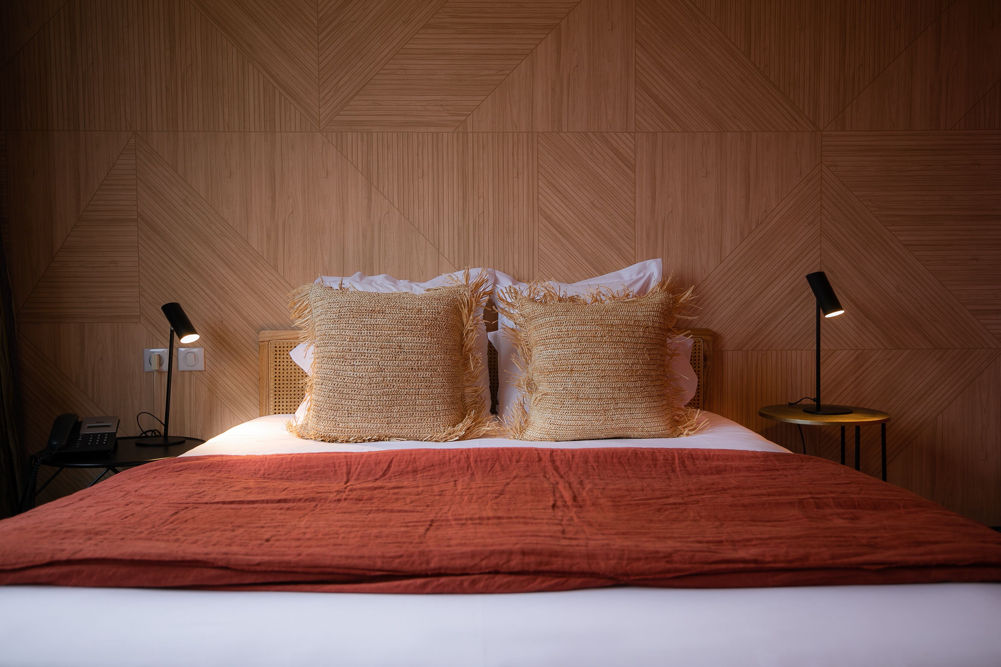 Ein Hotelzimmer mit einer Holzwand, einem Bett mit roter Decke, zwei strukturierten Kissen, einer Nachttischlampe und einem Telefon auf dem Nachttisch.