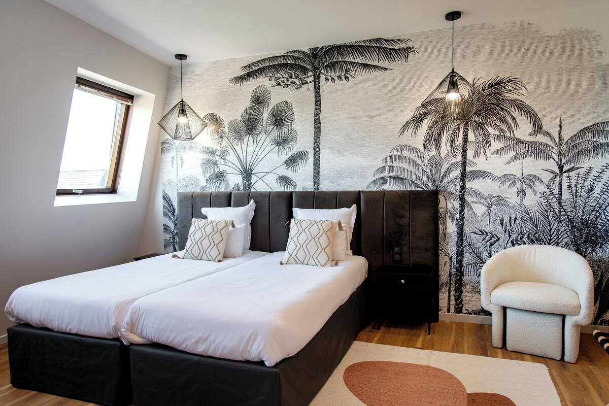 Een slaapkamer met twee bedden, een groot raam, een muurschildering van palmbomen en een moderne stoel.