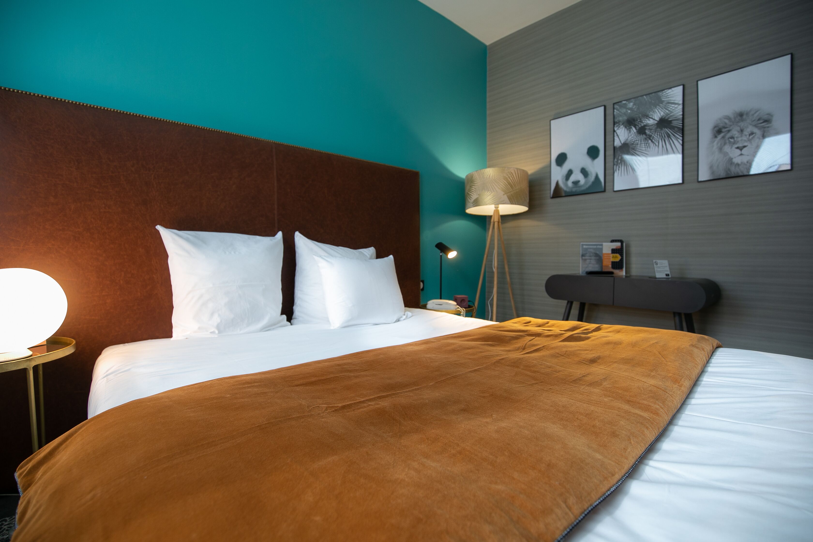 A hotel room with a large bed, a brown headboard, a white pillow, a brown throw blanket, a bedside lamp, and wall art including a panda and a lion.