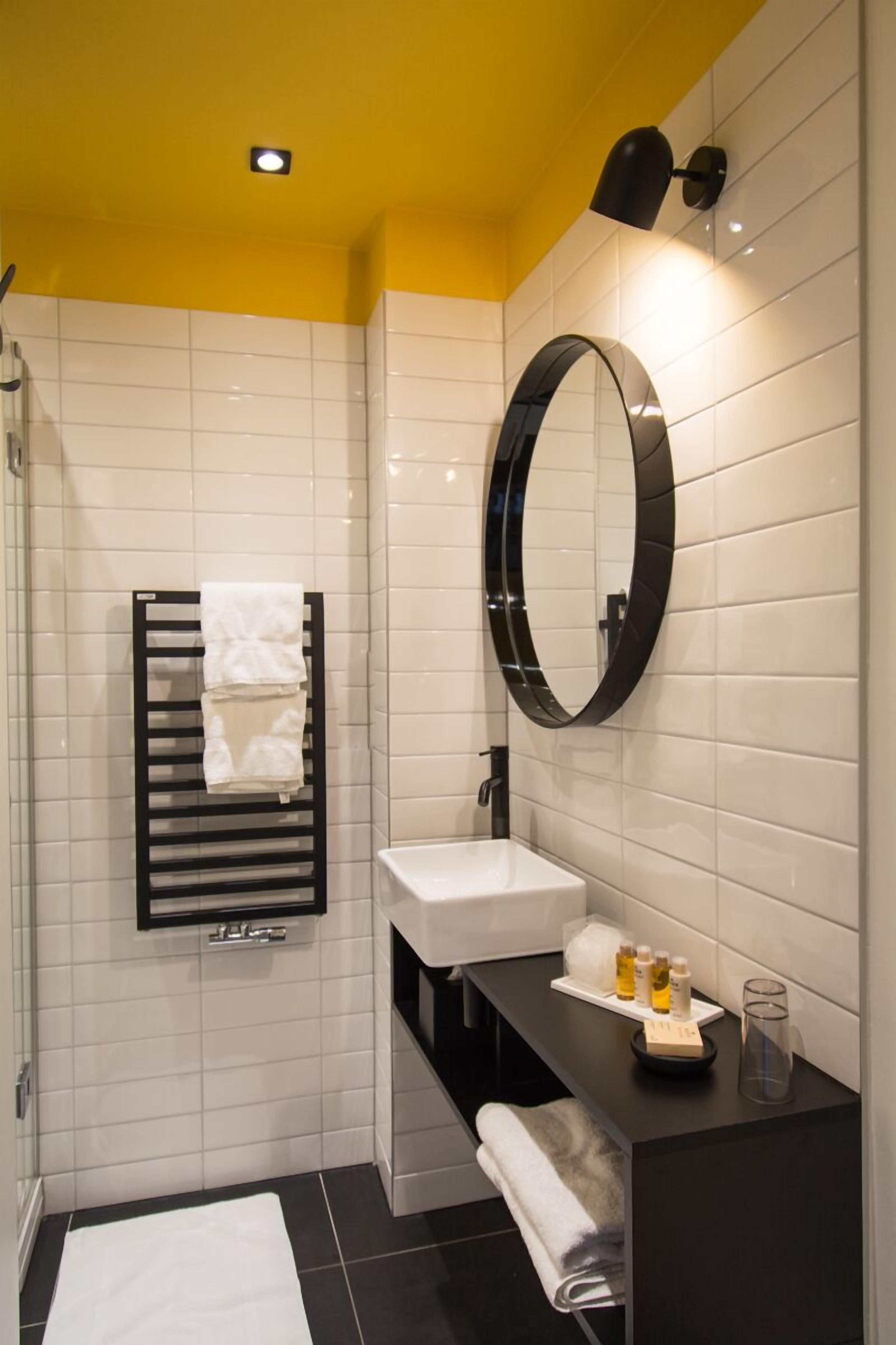 A modern bathroom with a white tiled wall, a black towel rack, a round mirror, a white sink, and a black countertop with toiletries.
