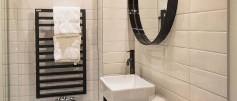 A modern bathroom with a white tiled wall, a black towel rack, a round mirror, a white sink, and a black countertop with toiletries.