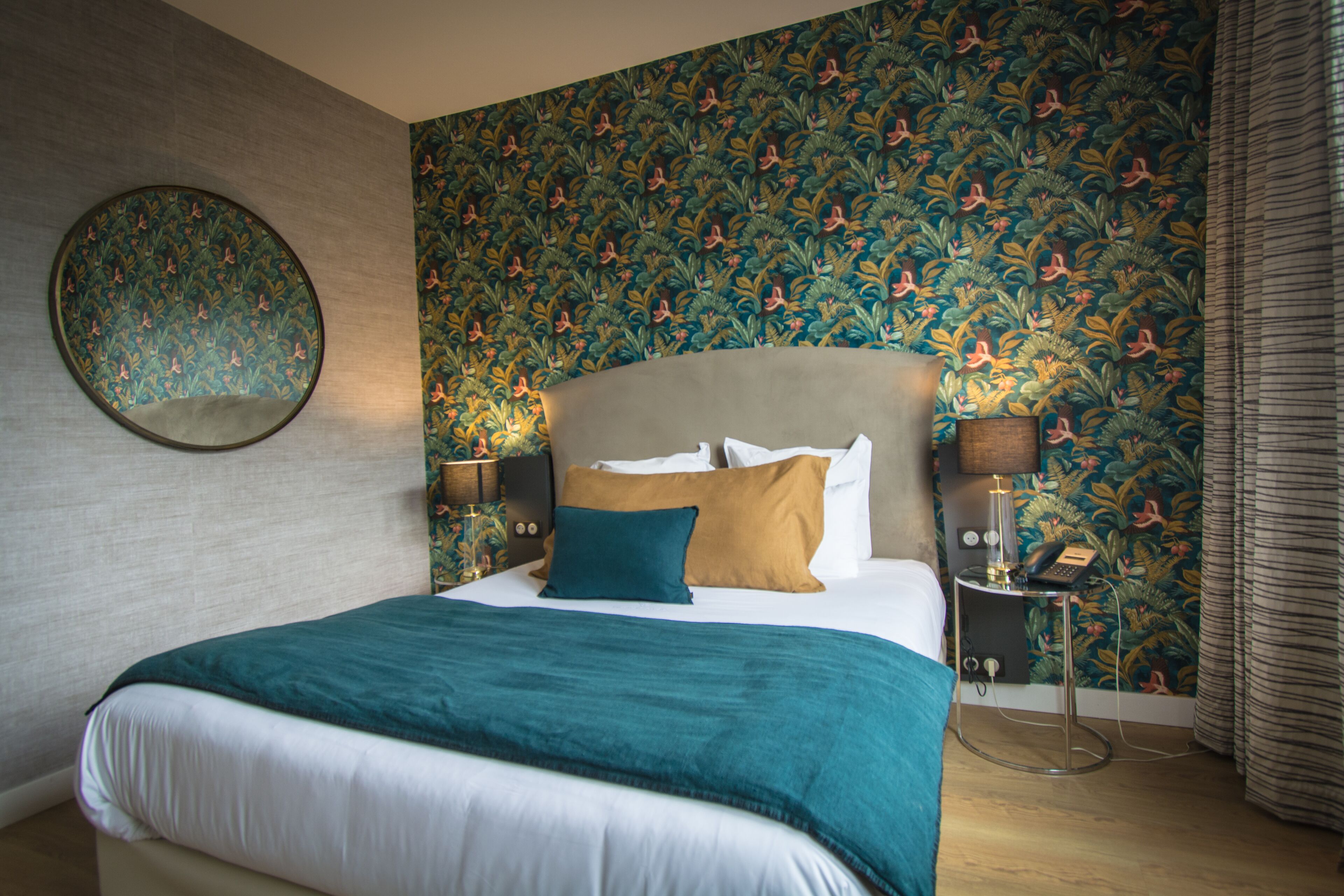 A neatly made bed with a teal blanket, a round mirror on the wall, and a floral wallpaper pattern.